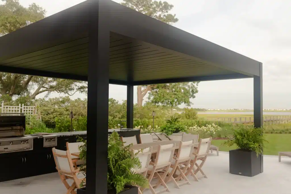 Black R-blade pergola covering an outdoor dining set and fully-equipped kitchen with a stainless steel grill, overlooking a marshy landscape.
