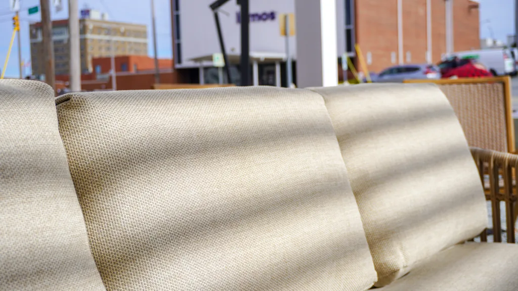 A close-up detail of the textured, light beige cushion of a high-end outdoor sofa at Broad Hall, with sunlight filtering through the slats of the R-Blade pergola creating striking shadow lines. The urban backdrop of High Point is blurred in the background.