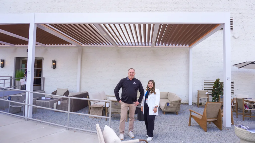 Two Azenco Outdoor representatives, a man and a woman, stand smiling beneath the dual-bay white and wood-look R-Blade motorized louvered pergola installation on the gravel patio at Broad Hall, showcasing the featured project at High Point Market.