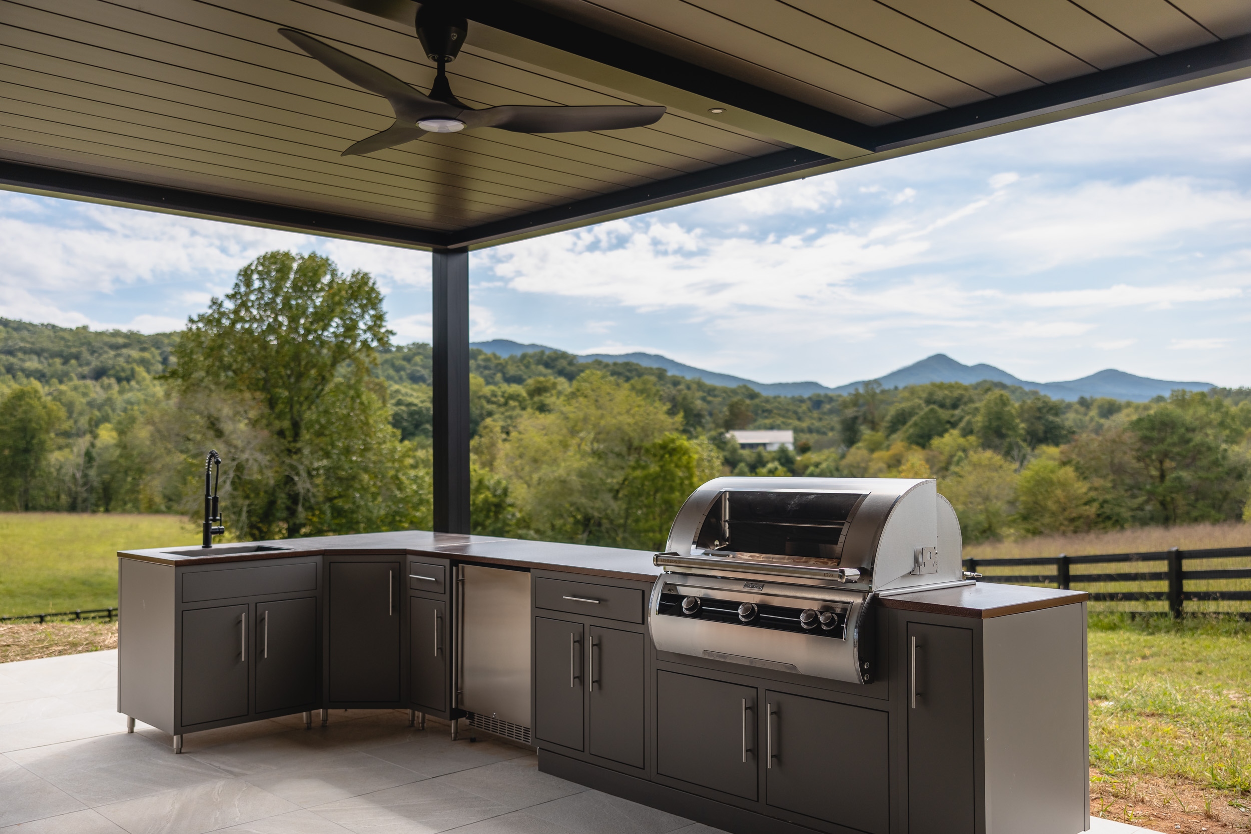 Black R-BLADE pergola with ceiling fans and LED lights over an outdoor kitchen with mountain views.