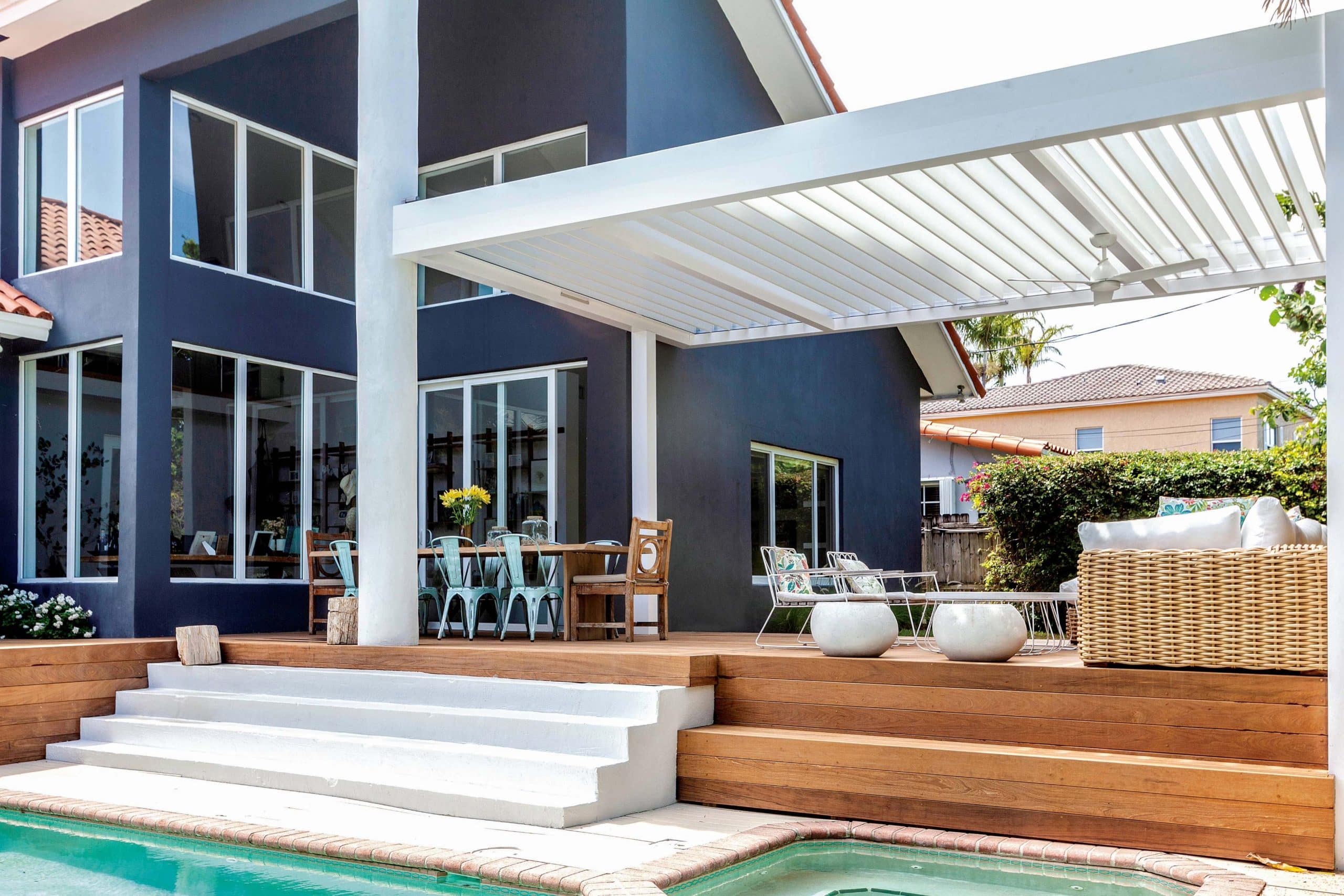 White R-BLADE pergola over a wooden deck beside a dark blue home and swimming pool.