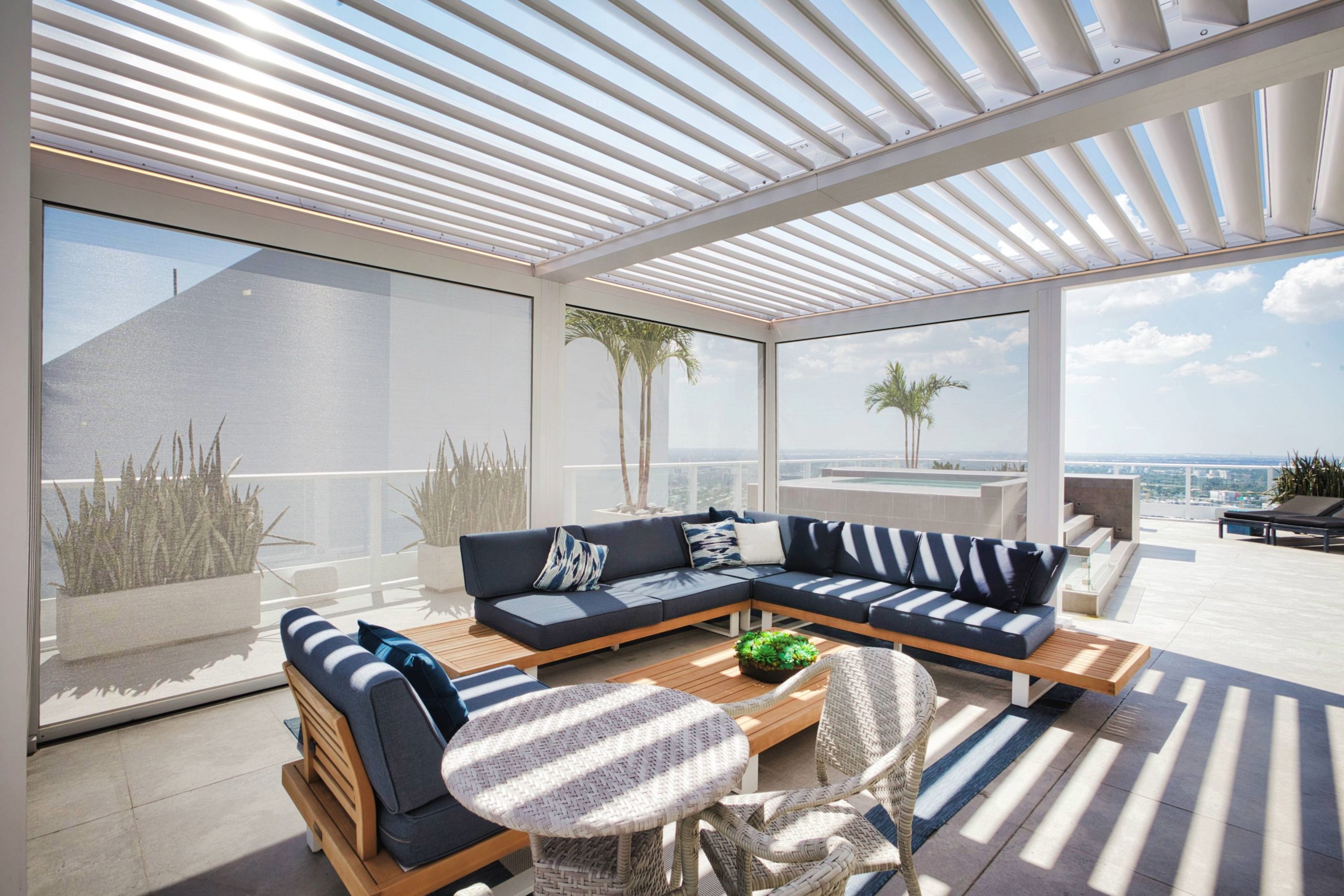 White louvered pergola shading a patio lounge with blue seating and an ocean view.
