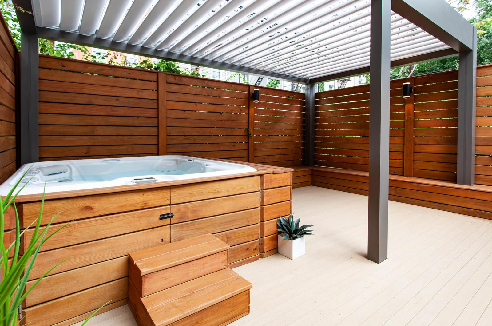 Hot tub beneath an R-BLADE pergola with white louvers and wooden privacy walls.